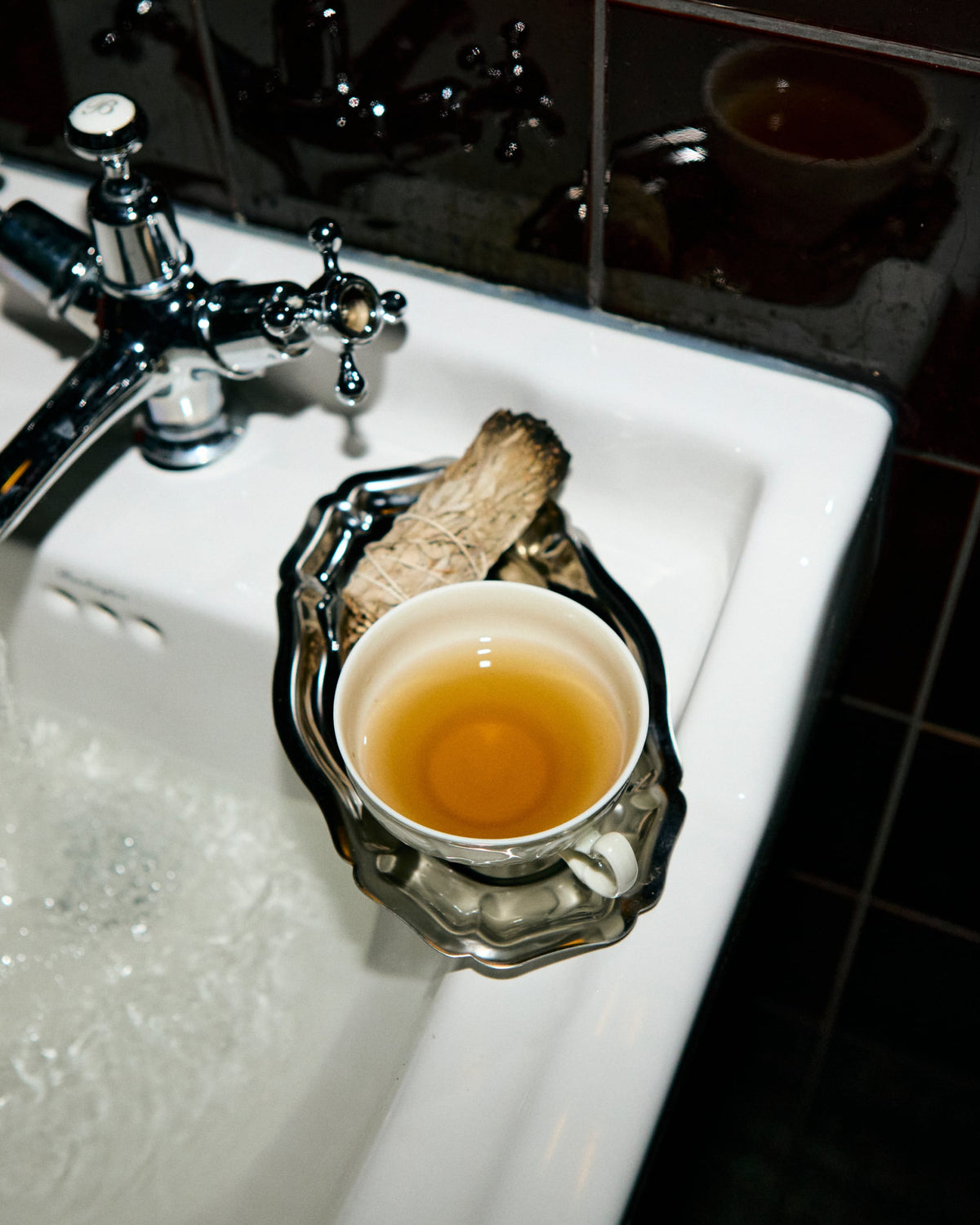Weiße Keramiktasse mit hellem Tee auf verziertem silbernem Unterteller am Waschbecken mit laufendem Wasserhahn.
