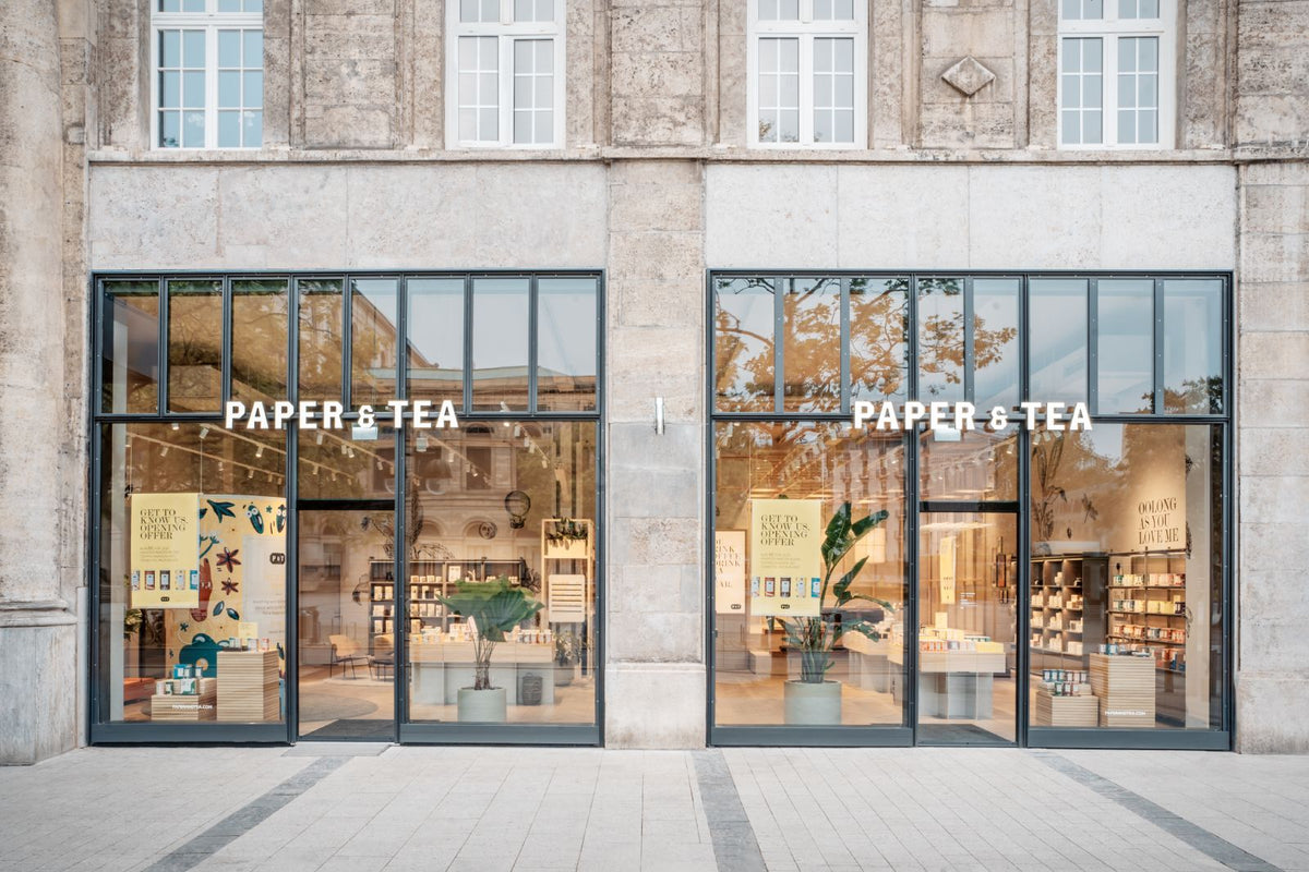 Moderner Teeladen PAPER & TEA mit gläsernen Eingängen, beleuchtet, mit Teeprodukten und Werbeplakaten.