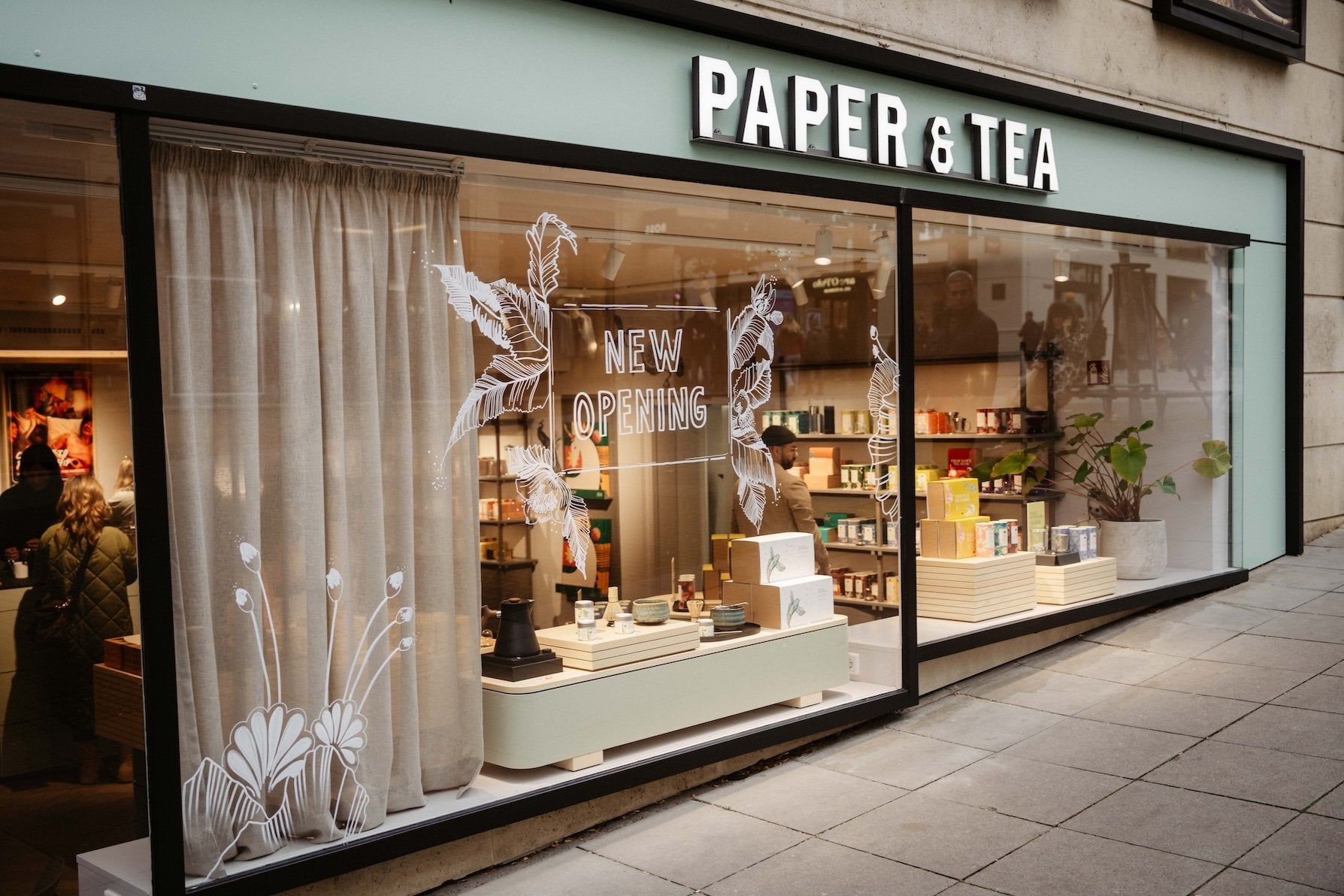 PAPER & TEA Schaufenster mit 'NEW OPENING', floralen weißen Illustrationen, Teeprodukten und Kunden im Innenraum.