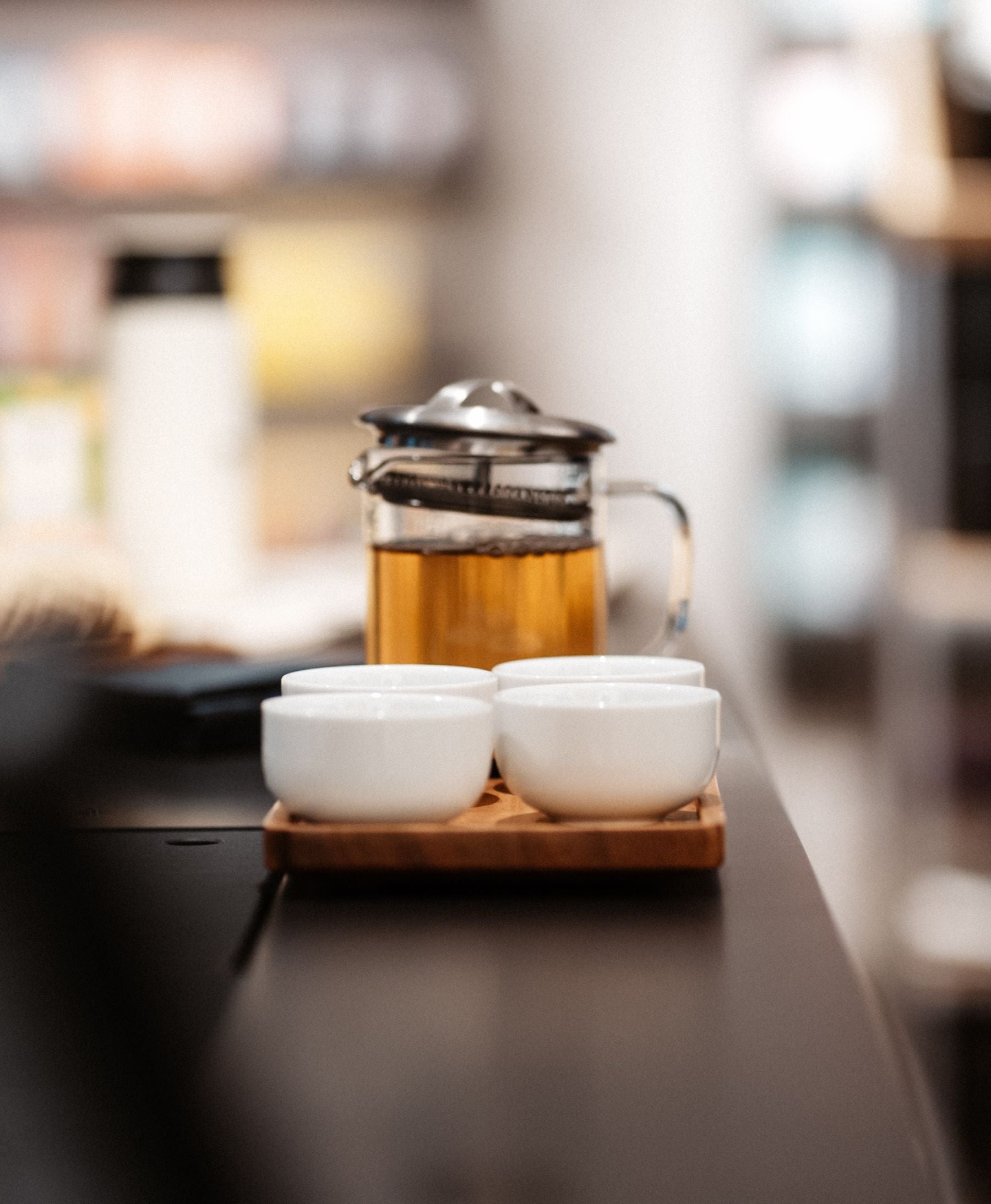 Glas-French-Press mit bernsteinfarbenem Tee und vier weißen Keramiktassen auf Holztablett im warmen Licht.