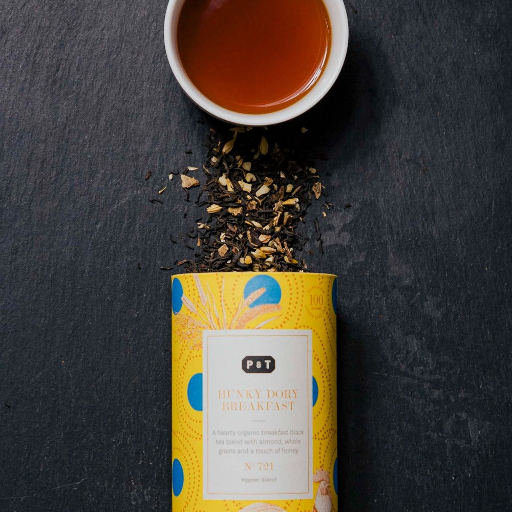 Gelbe P & T HUNKY DORY BREAKFAST Teedose mit schwarzen Teeblättern, Mandeln und Getreide neben Tasse bernsteinfarbenem Bio-Schwarztee.