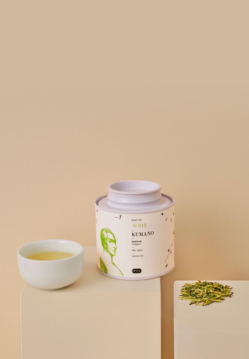 Beige Teedose mit KUMANO KUKICHA Karigane BIO Grüntee, daneben Tasse mit grünem Tee und Teeblättern.