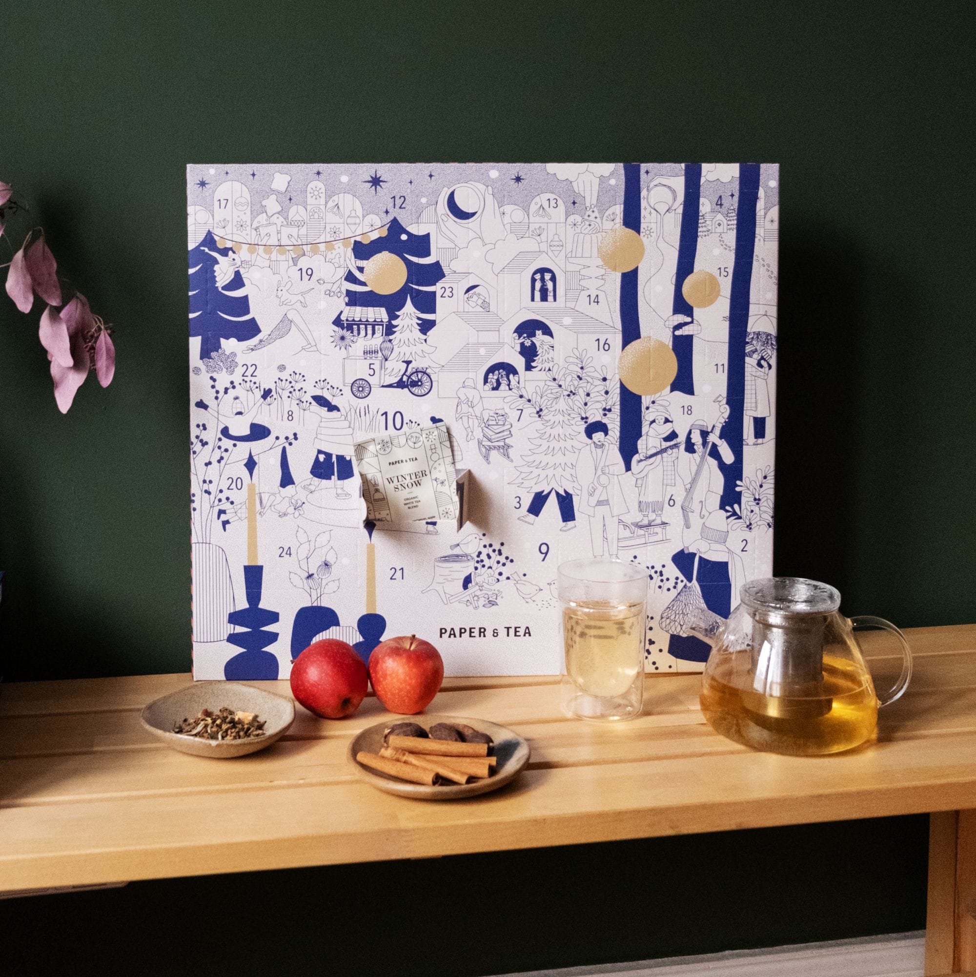 Festlicher Papier-Adventskalender von PAPER & TEA auf Tisch mit Teekanne, Tasse, Äpfeln und Zutaten.