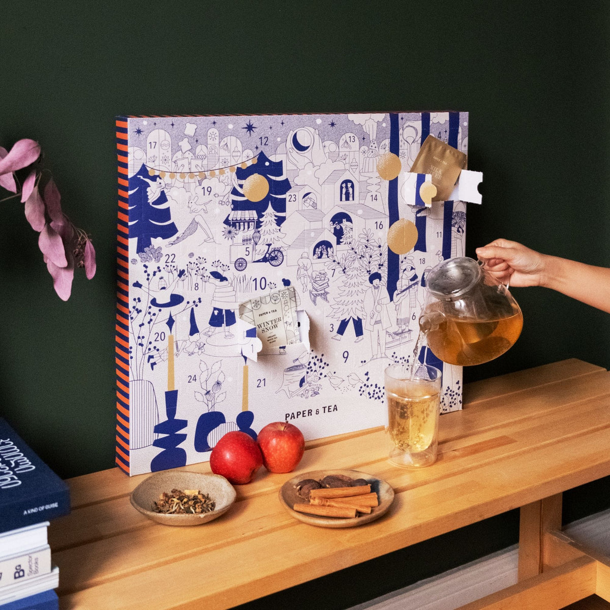 Adventskalender 'Winter Snow' von Paper & Tea mit blauen und goldenen Illustrationen, Teekanne gießt Tee ein, Äpfel, Zimt und Schokolade auf Holzbank.