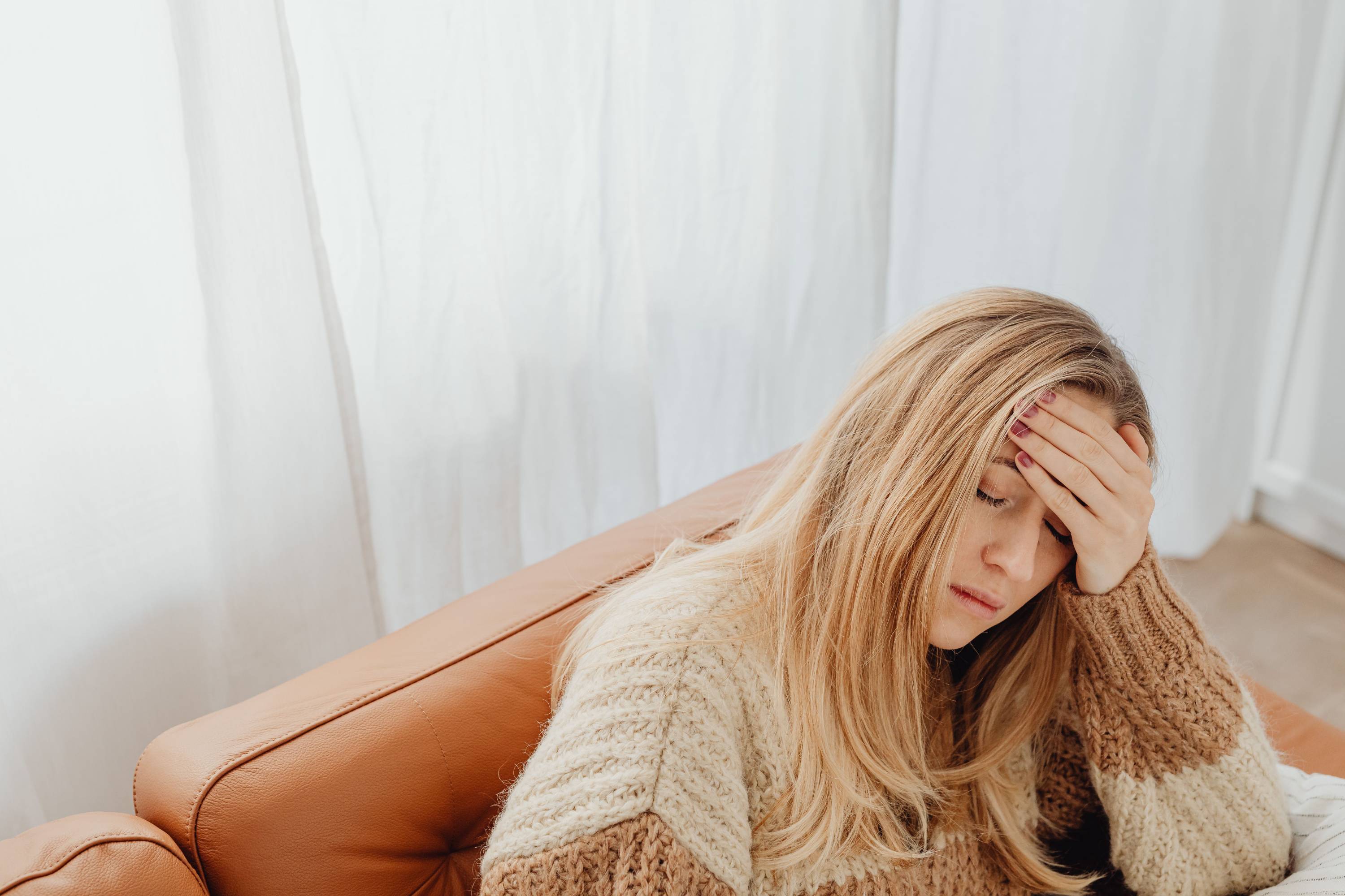 Junge blonde Frau im gestreiften Strickpullover auf Ledersofa hält sich die Hand an den Kopf