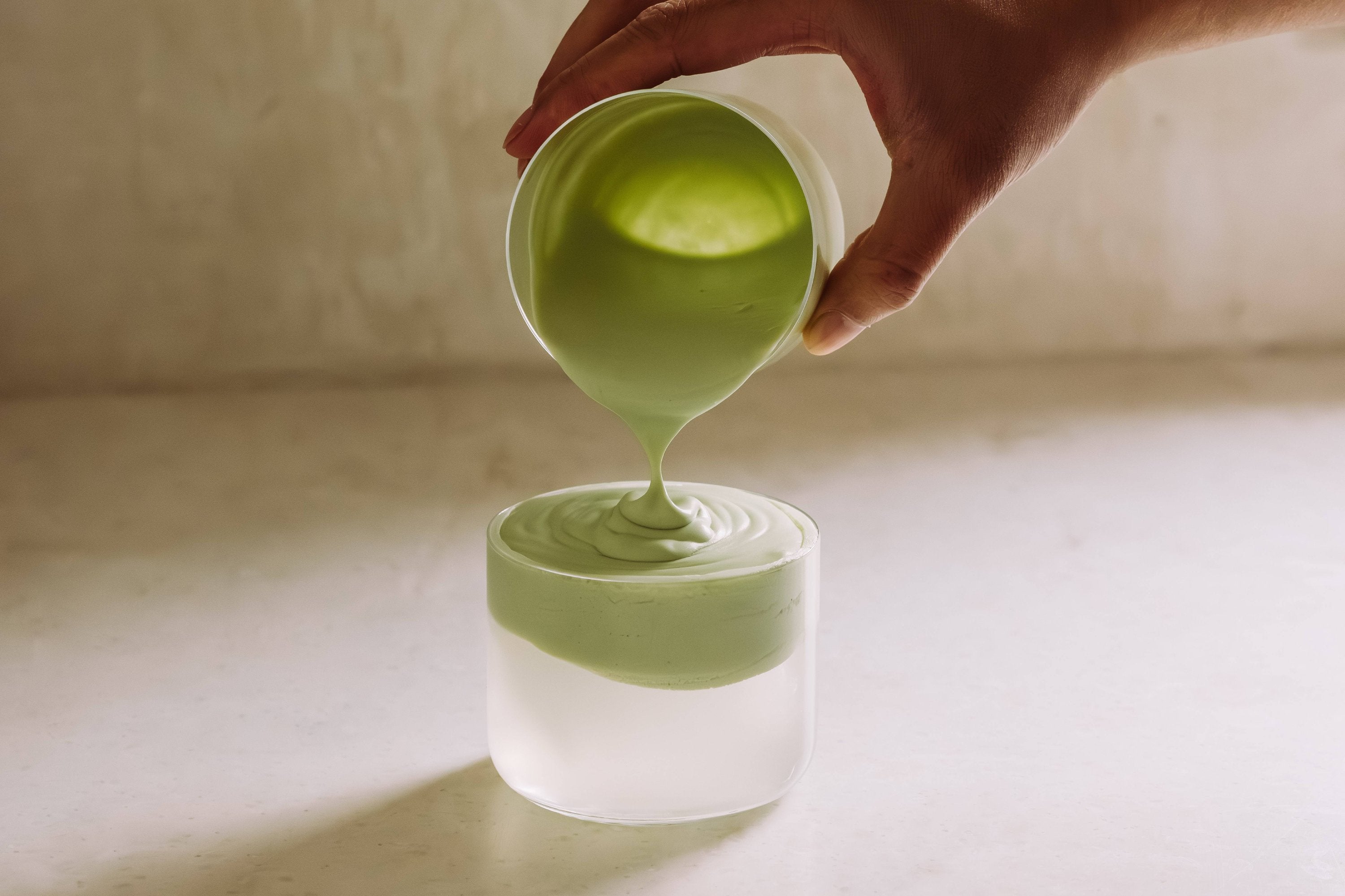 Eine Hand gießt blassgrüne Matcha-Creme in Glas