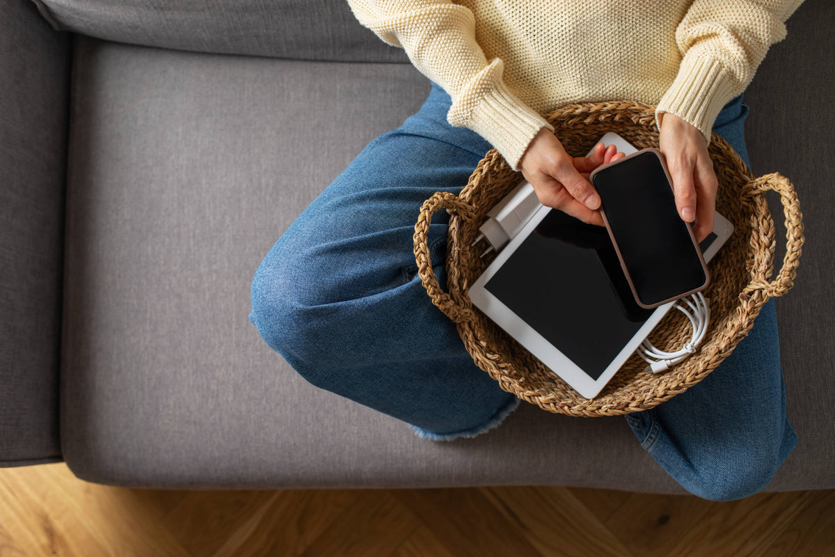 Person auf grauem Sofa mit blauer Jeans und Strickpullover, gibt Smartphone in einen Korb mit Tablet