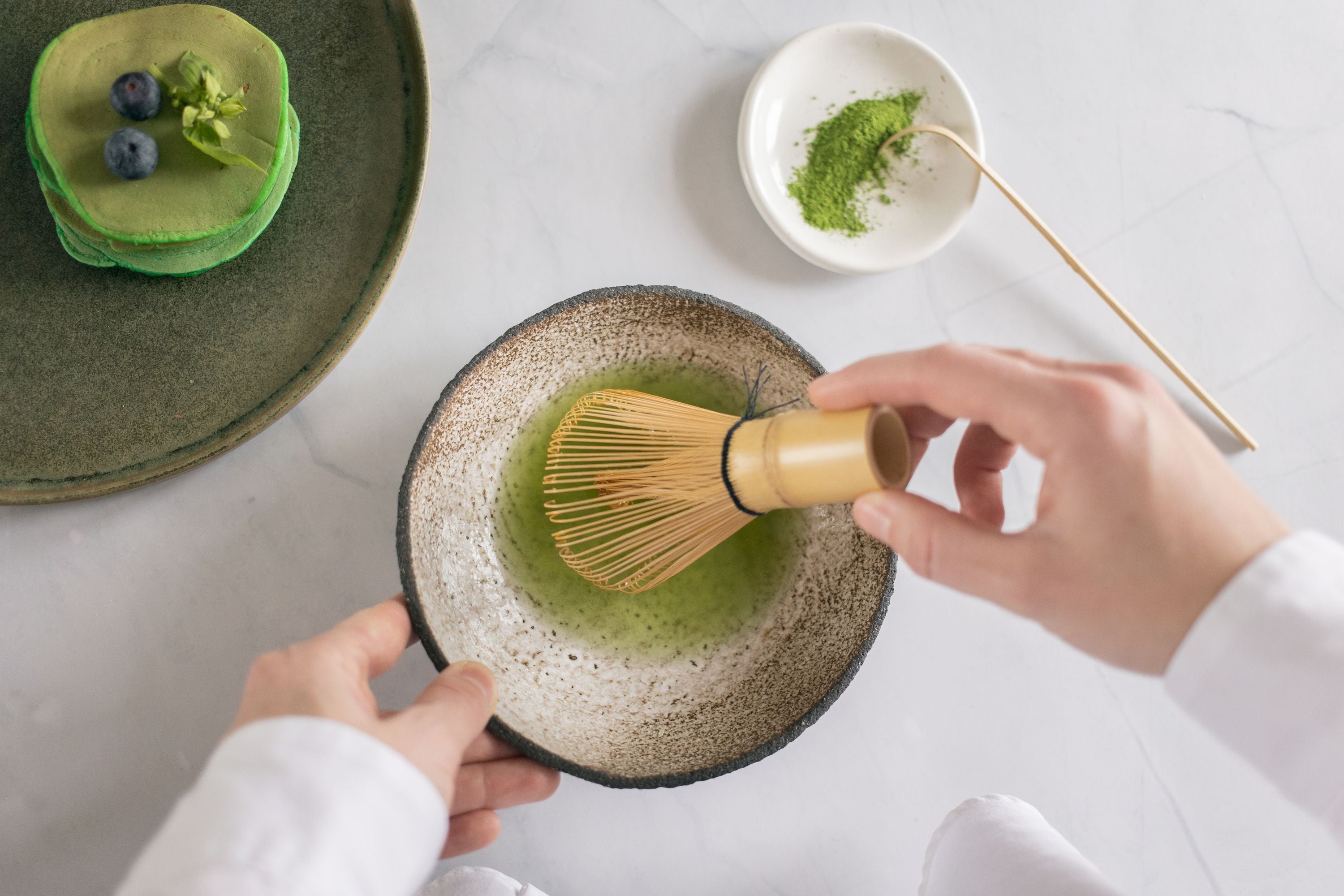 Hände schlagen lebendigen grünen Matcha-Tee mit Bambuschasen in rustikaler Keramikschale auf, daneben Matcha-Pfannkuchen mit Heidelbeeren
