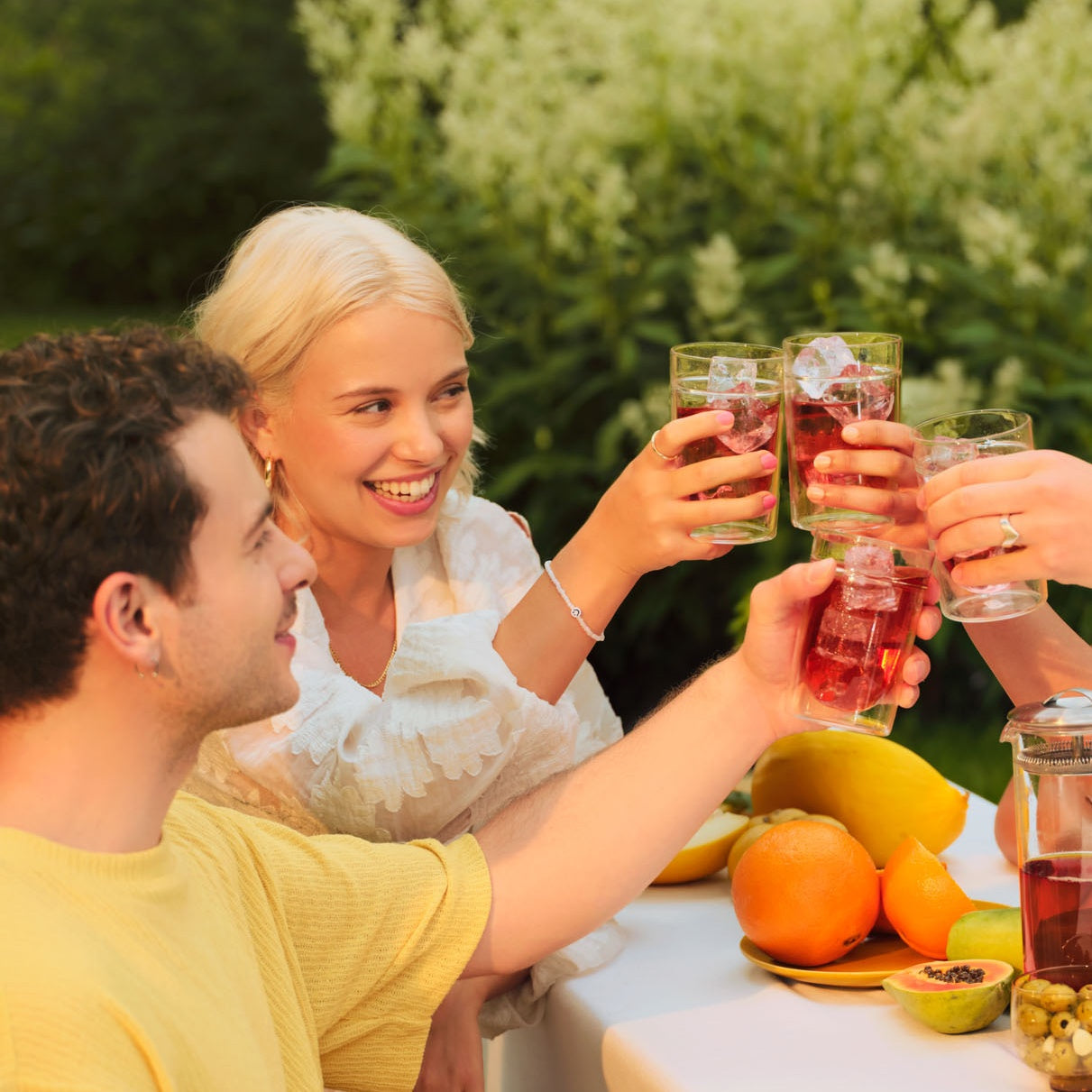Drei junge Menschen stoßen mit Gläsern voller rotem Getränk an, umgeben von Orangen und Papayas auf einem weißen Tisch im Freien.
