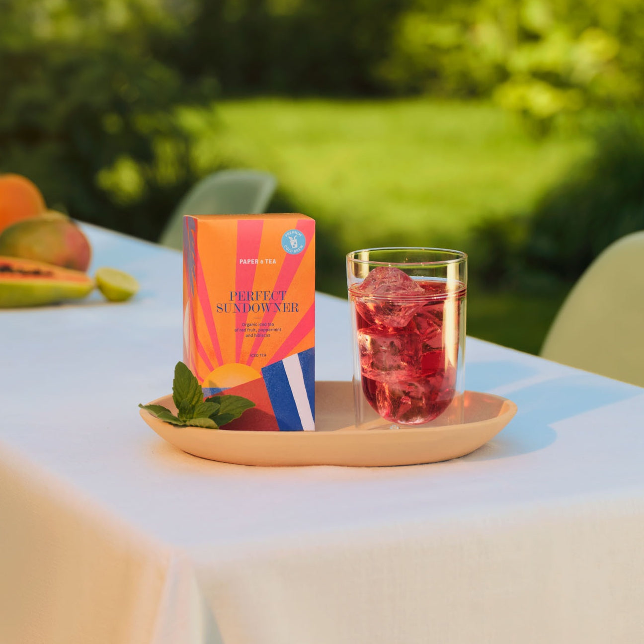 PAPER & TEA Perfect Sundowner Bio-Eistee mit roten Früchten, Pfefferminze und Hibiskus auf Tablett im Garten.