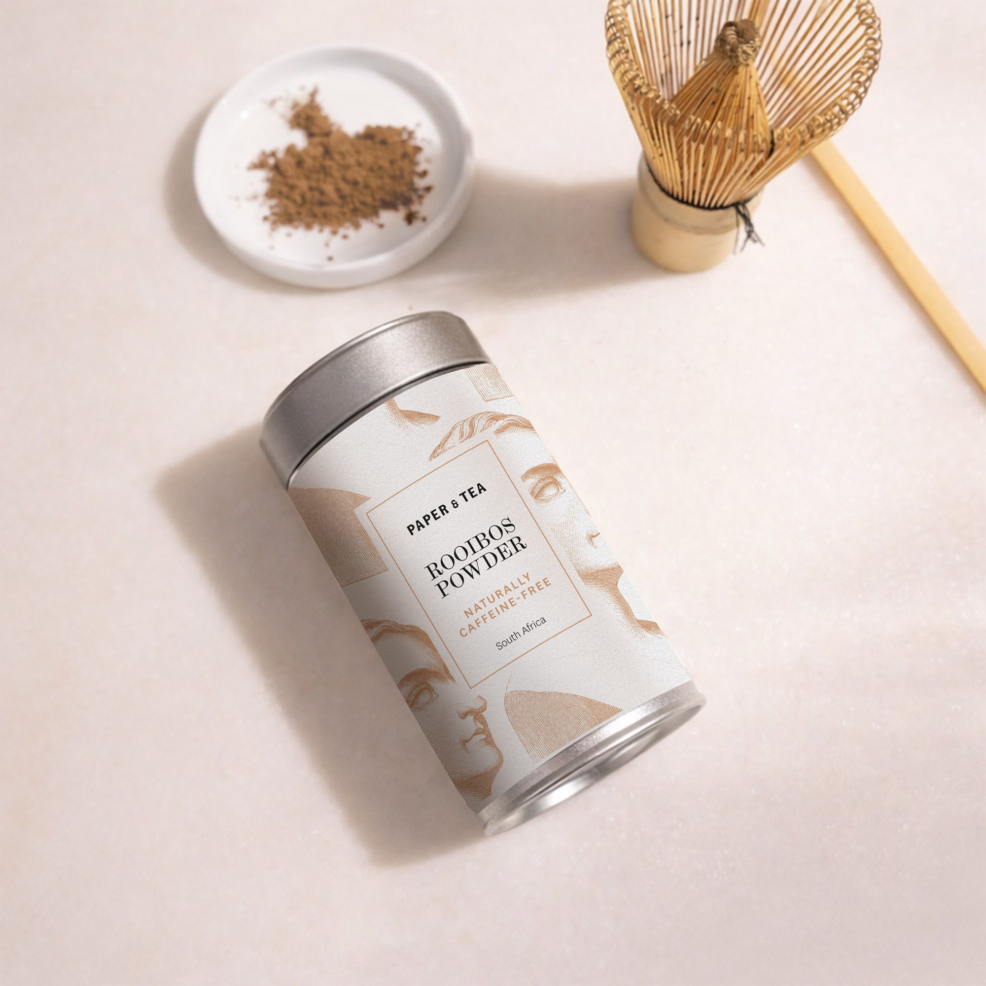 Elegante Anordnung mit Rooibos-Pulver in Metalldose und Schale, abgebildet mit einem Bambusbesen.