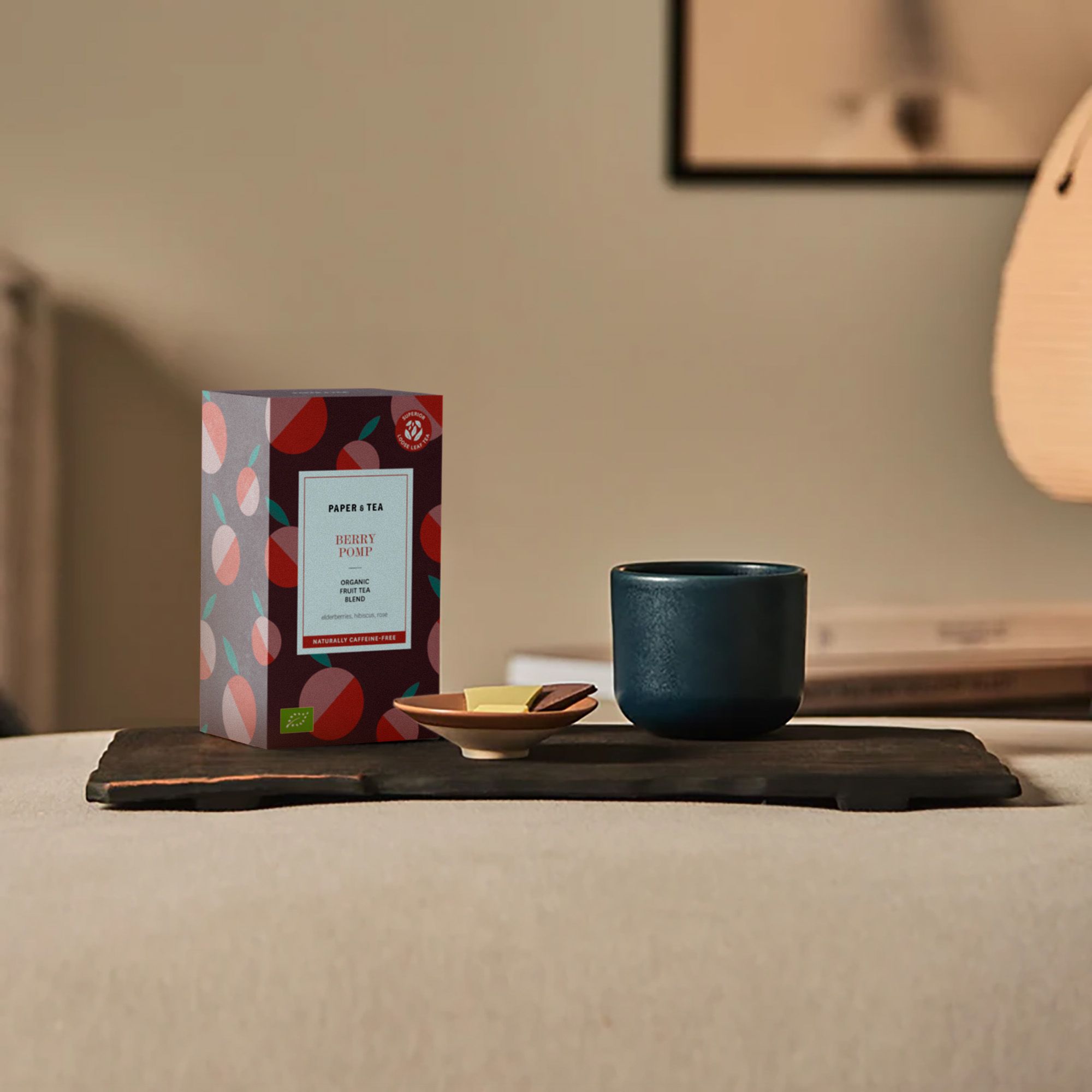 Bunte Box mit PAPER & TEA BERRY POMP Bio-Früchtetee, Schokoladenstücken und Tasse auf Holzbrett.