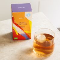 Bunte SANDY SWEET JOY Verpackung, Erdnussbutter-Banane-Honig-Snack neben Glas mit Getränk auf Marmortisch.