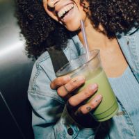 Junge Frau mit lockigem Haar lächelt und genießt grünen Tee in gemütlichem Caféambiente.