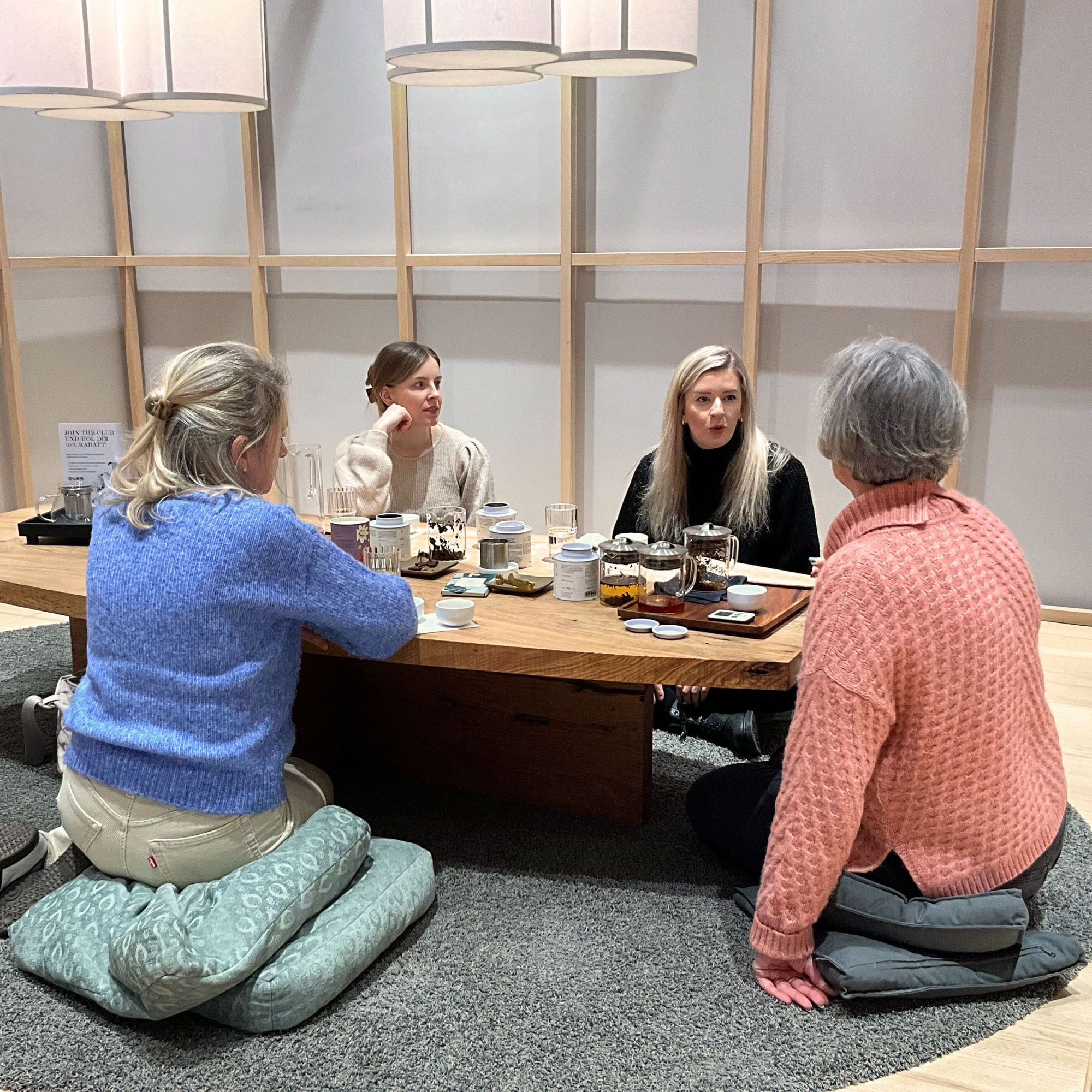 Vier Frauen sitzen auf Bodenkissen an einem niedrigen Holztisch, trinken Tee und unterhalten sich entspannt.