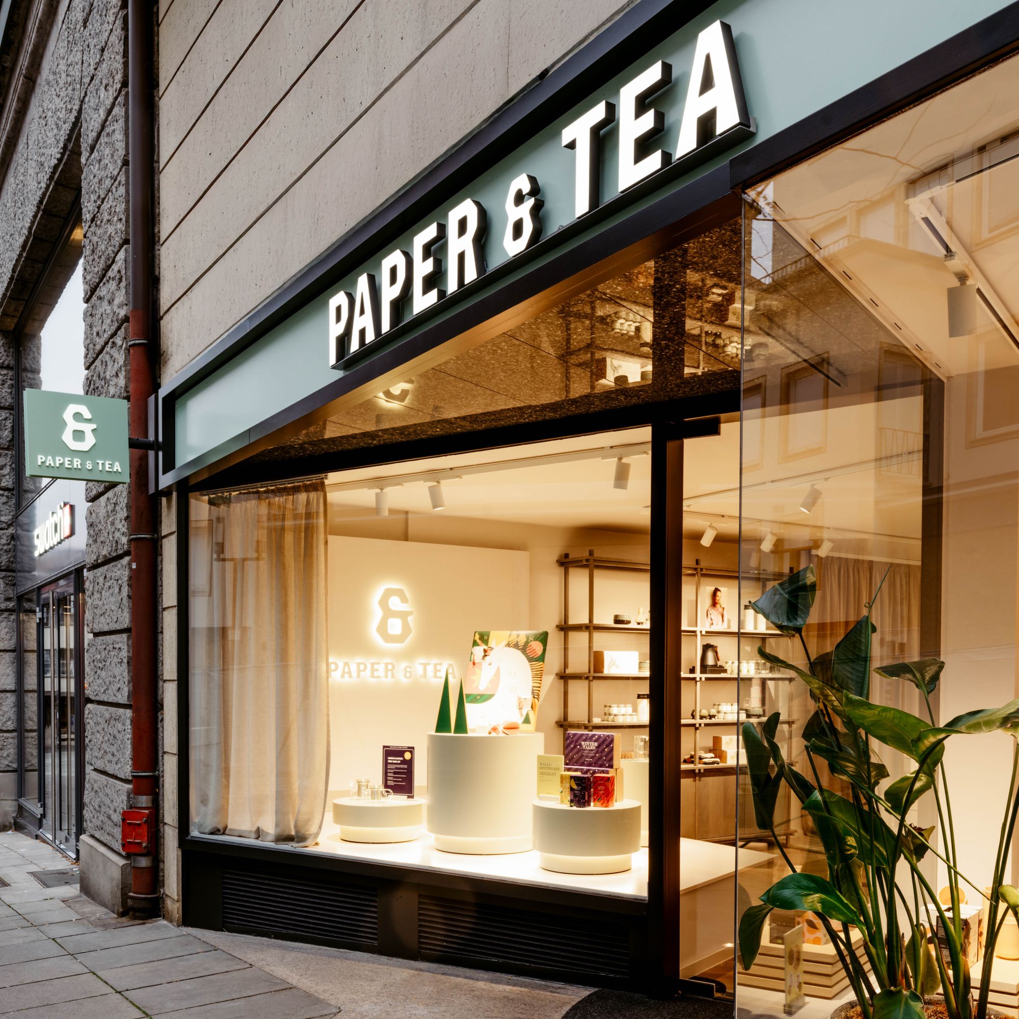 Moderne PAPER & TEA Ladenfassade mit beleuchtetem Logo und Schaufensterdekoration.