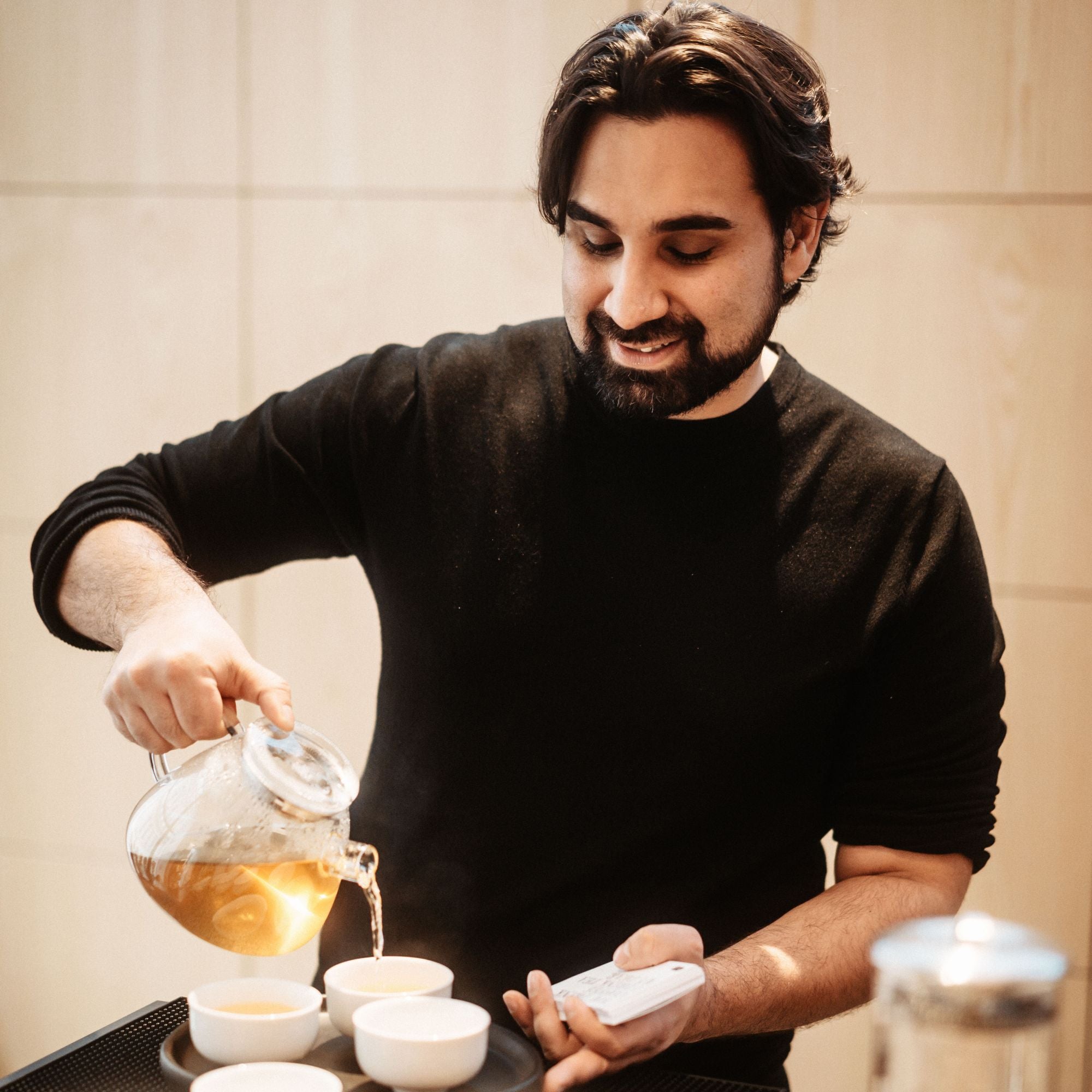 Mann mit dunklem Haar gießt dampfenden Tee aus Glasteekanne in weiße Tassen auf schwarzem Tablett.