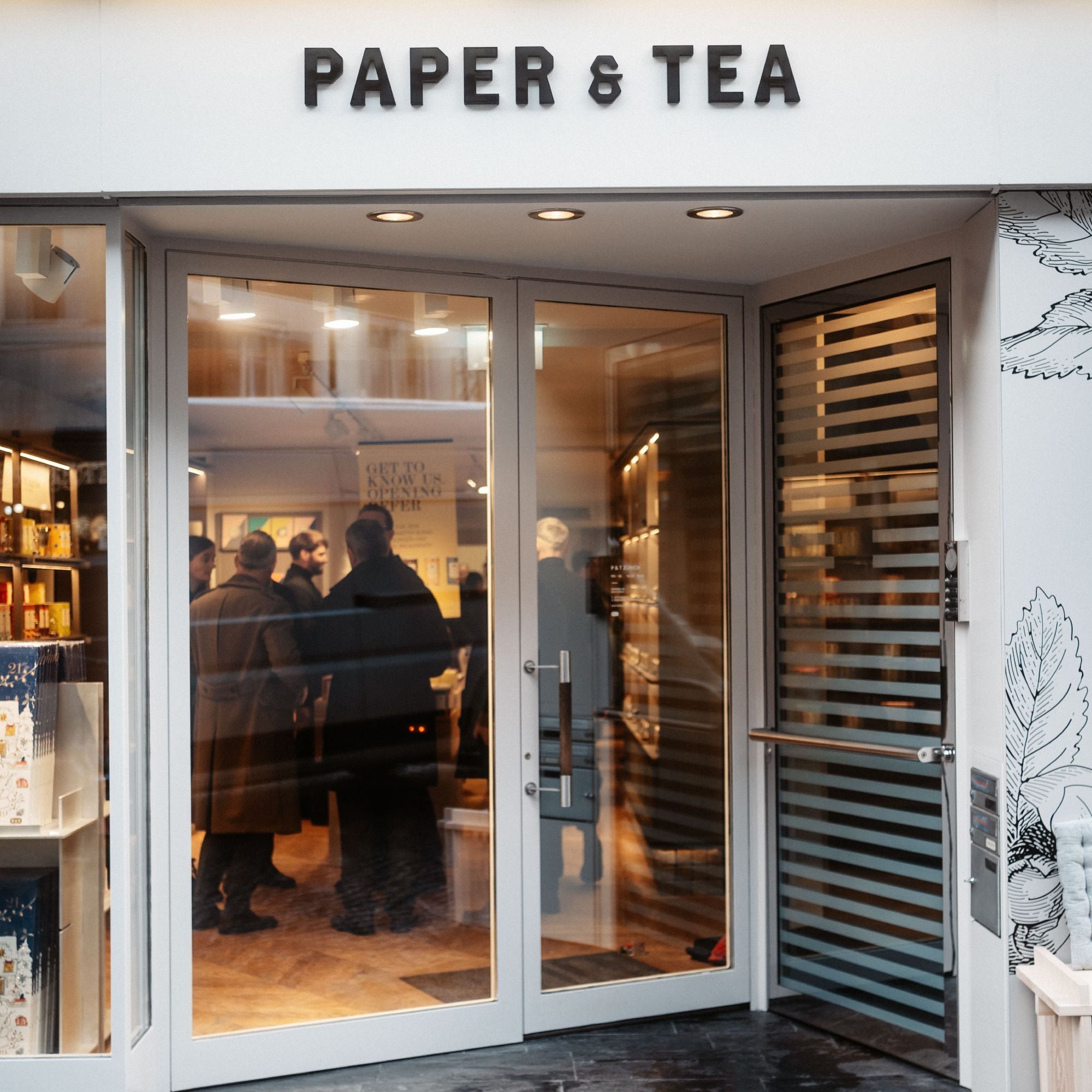 Eingang zum PAPER & TEA Geschäft mit Glas-Doppeltüren und blattförmigen Illustrationen an der Wand.