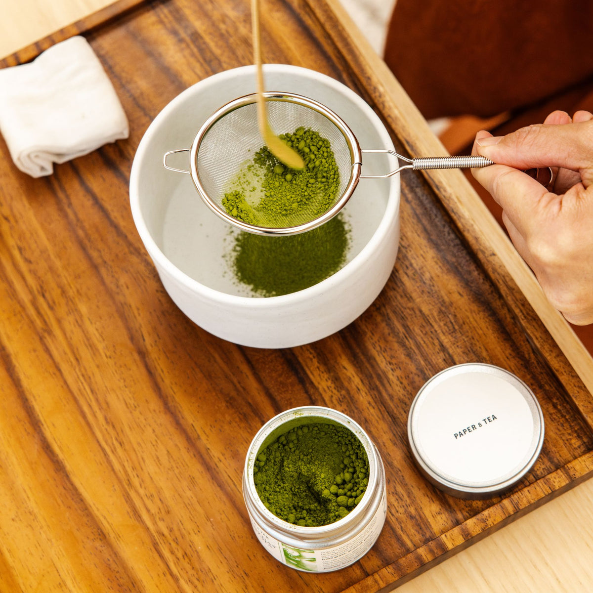 Matcha-Pulver wird mit einem Bambuslöffel in eine Keramikschale gesiebt, daneben Metall-Döschen auf Holztablett.
