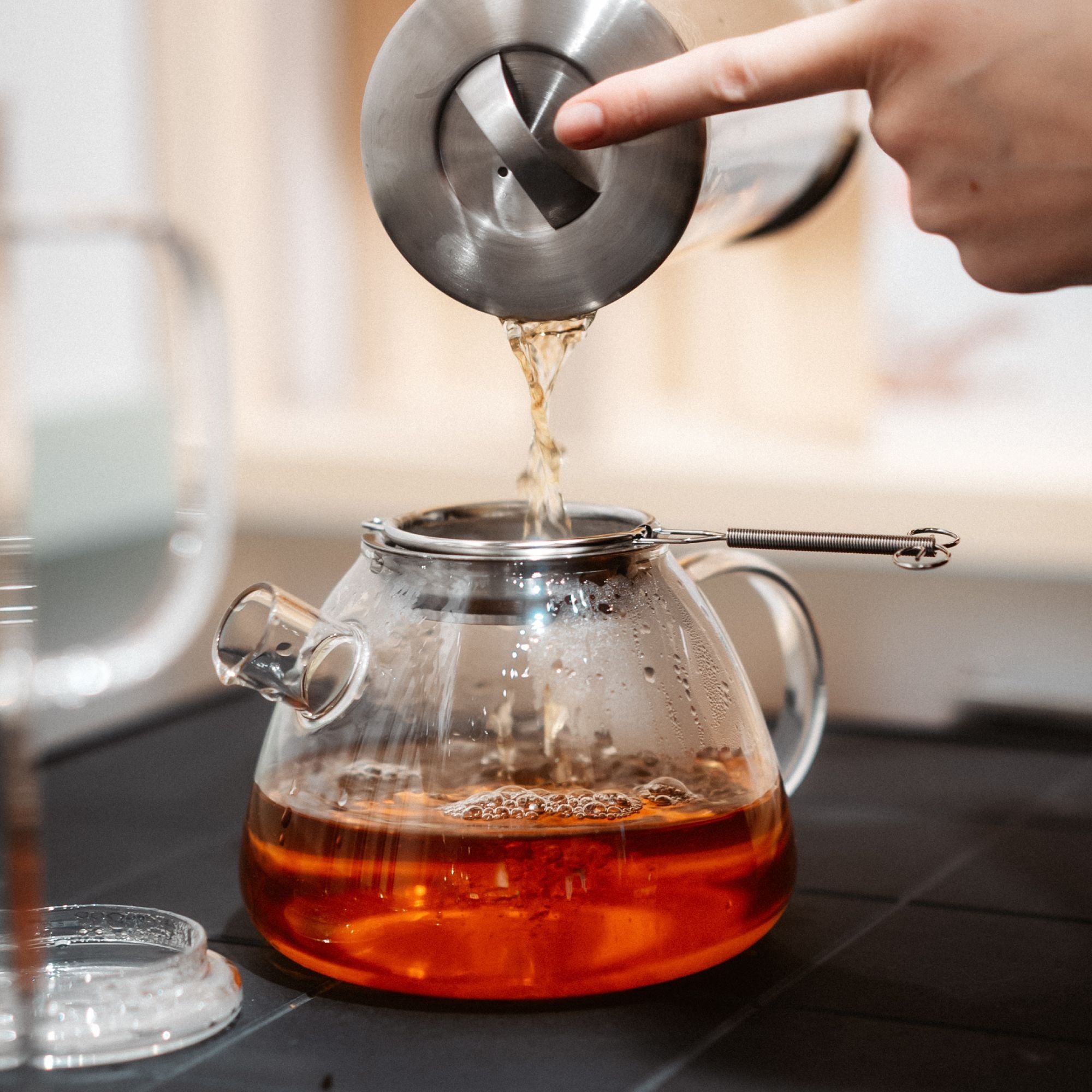 Glasteekanne mit Metallsieb, heißer Tee aus Kessel wird eingegossen, auf dunklem Hintergrund