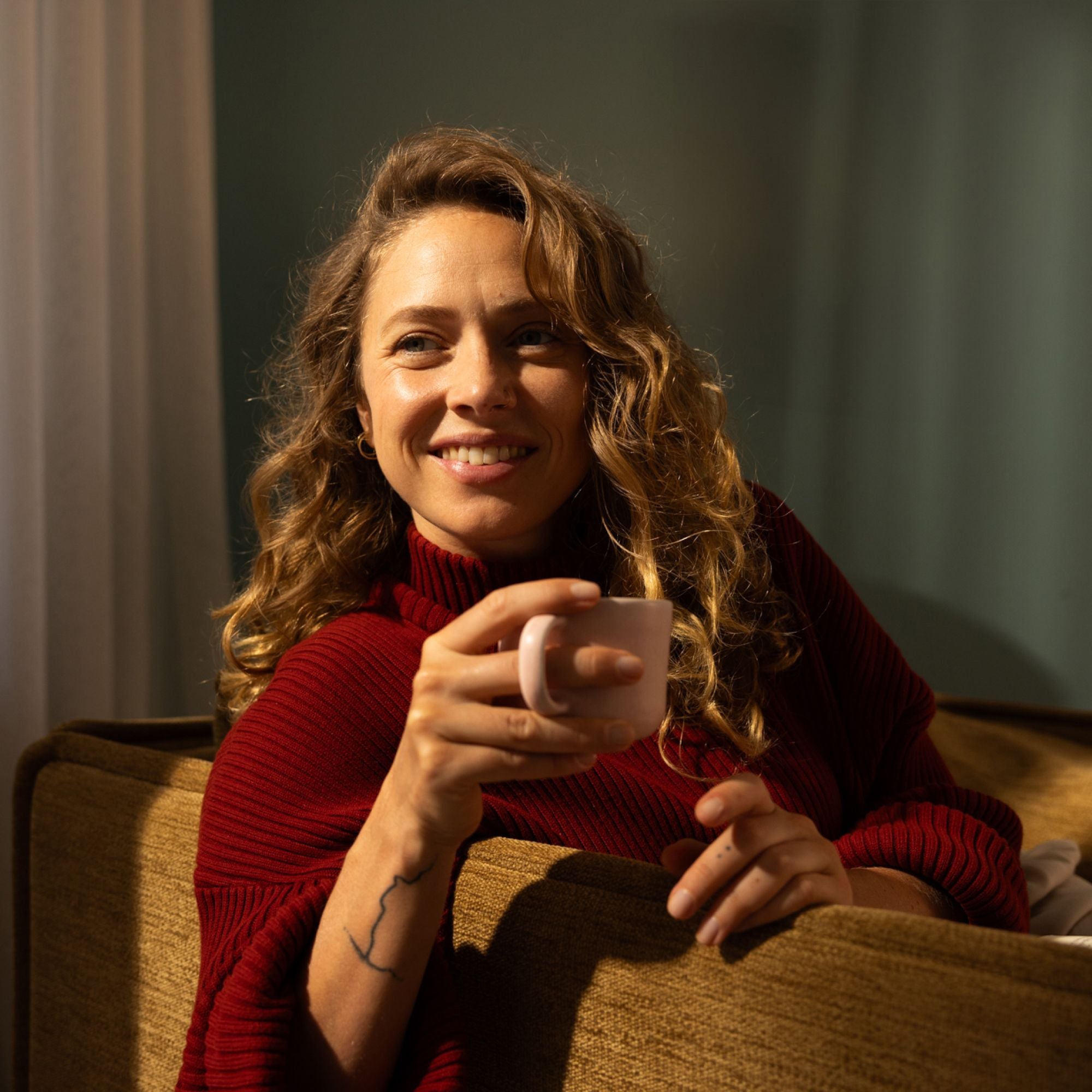 Lächelnde Frau mit lockigem Haar in rotem Pullover hält rosa Tasse auf braunem Sofa.