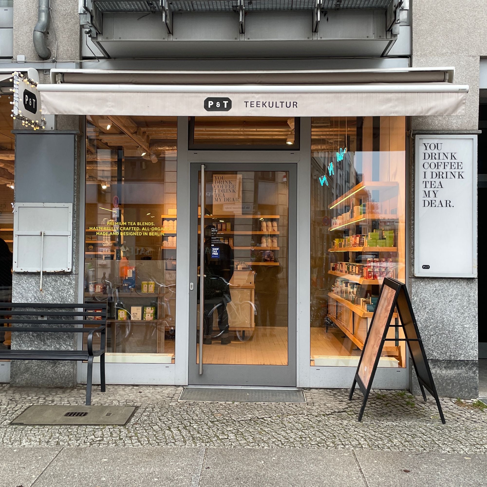 P & T TEEKULTUR Teehaus mit beleuchteten Regalen und 'YOU DRINK COFFEE I DRINK TEA MY DEAR.' Schriftzug.