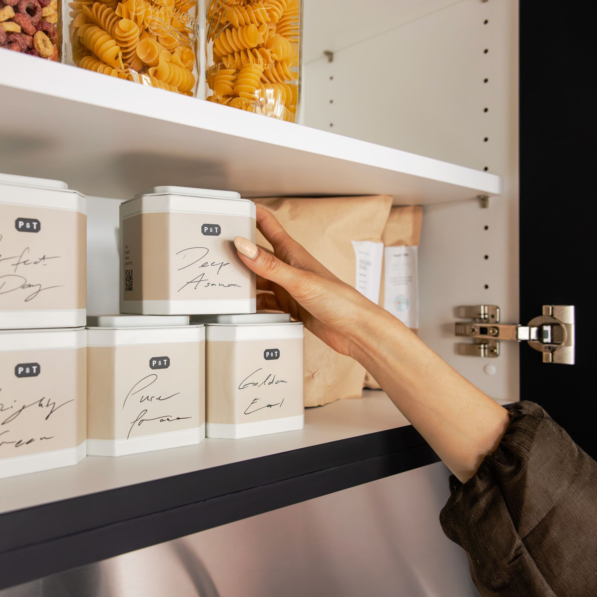 Hand mit gepflegten Nägeln greift nach beige P&T Teedose im Küchenschrank mit Teedosen und Pasta.