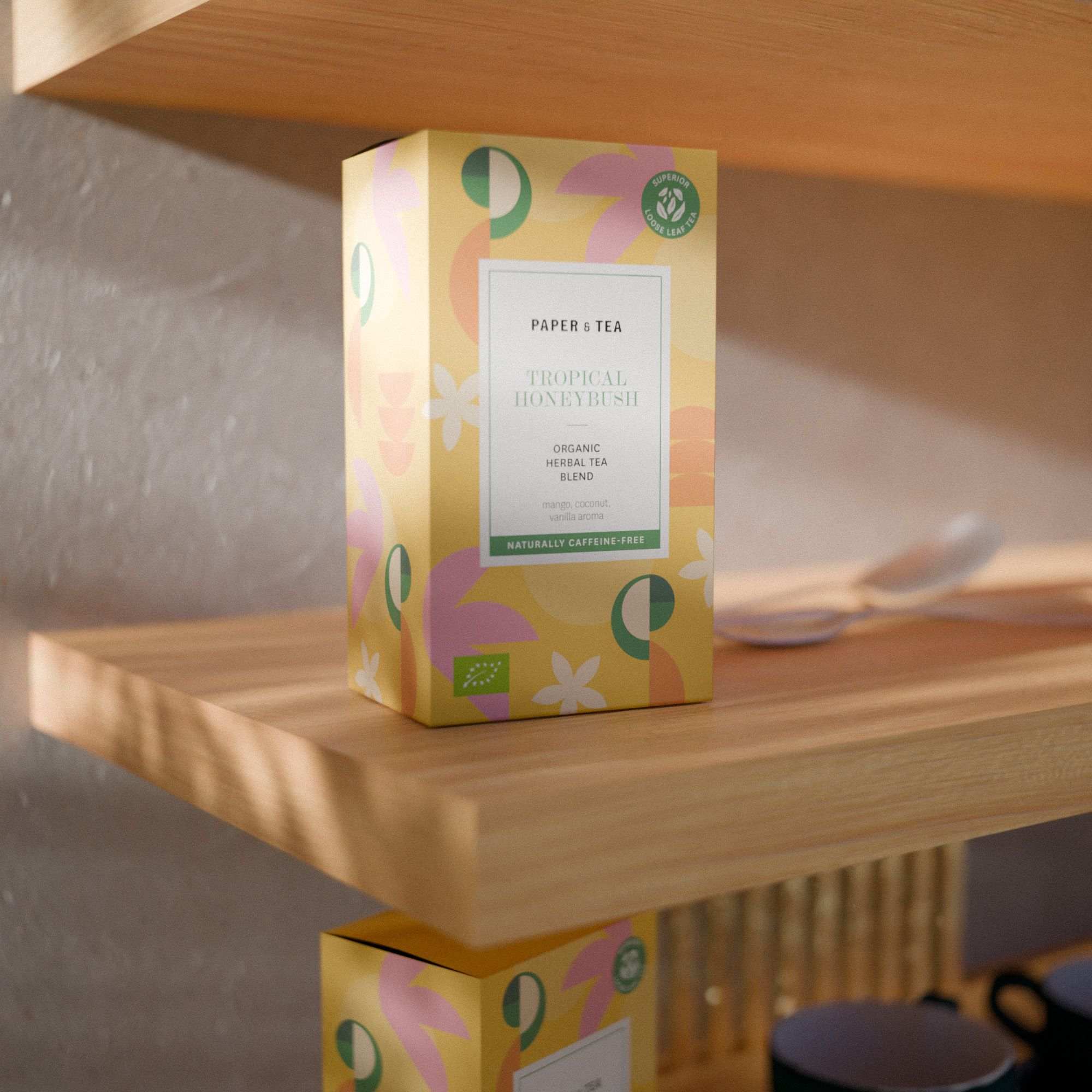 Bunte Packung Paper & Tea Tropical Honeybush Tee mit Mango-Kokos-Vanille Aroma auf Holzregal.