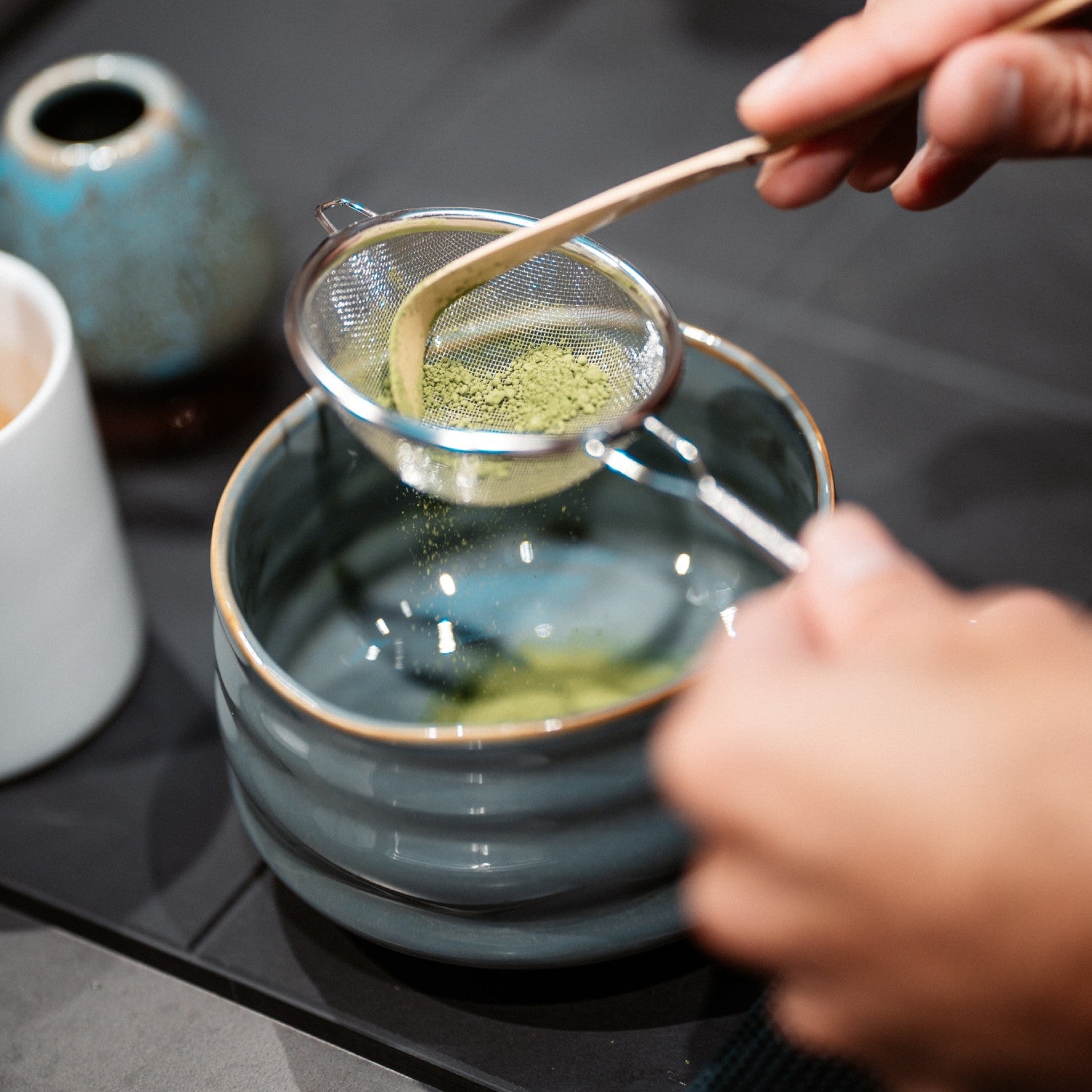Matcha-Pulver wird durch ein Sieb in eine Keramikschüssel gesiebt, Hände mit Bambuslöffel.