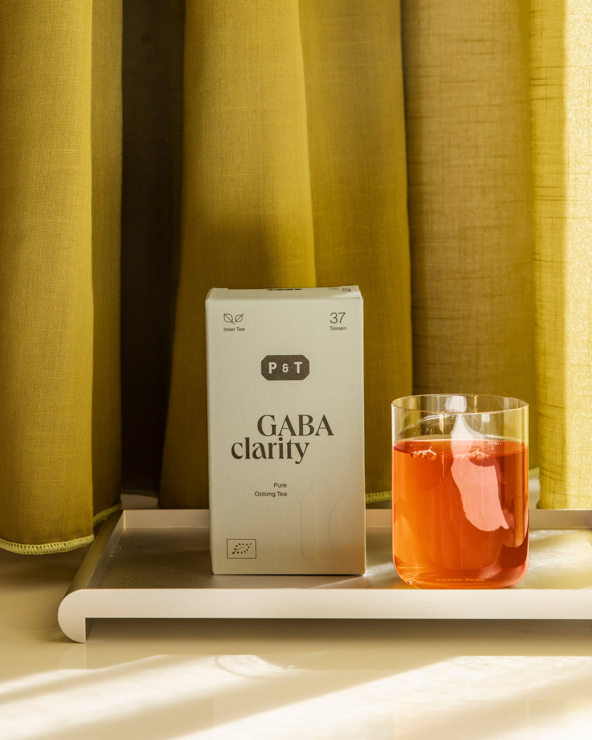 P&T GABA Clarity Oolong-Tee neben Glastasse mit rötlich-orangem Tee auf silbernem Tablett vor Vorhängen.