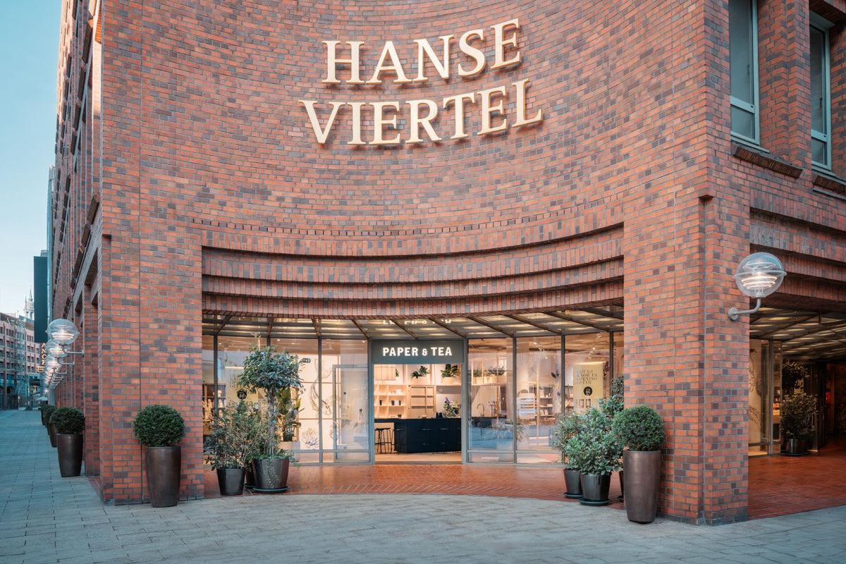 Eingang des P&T Ladens im Hanse Viertel mit goldenen Buchstaben und symmetrischen Pflanzen am Backstein.