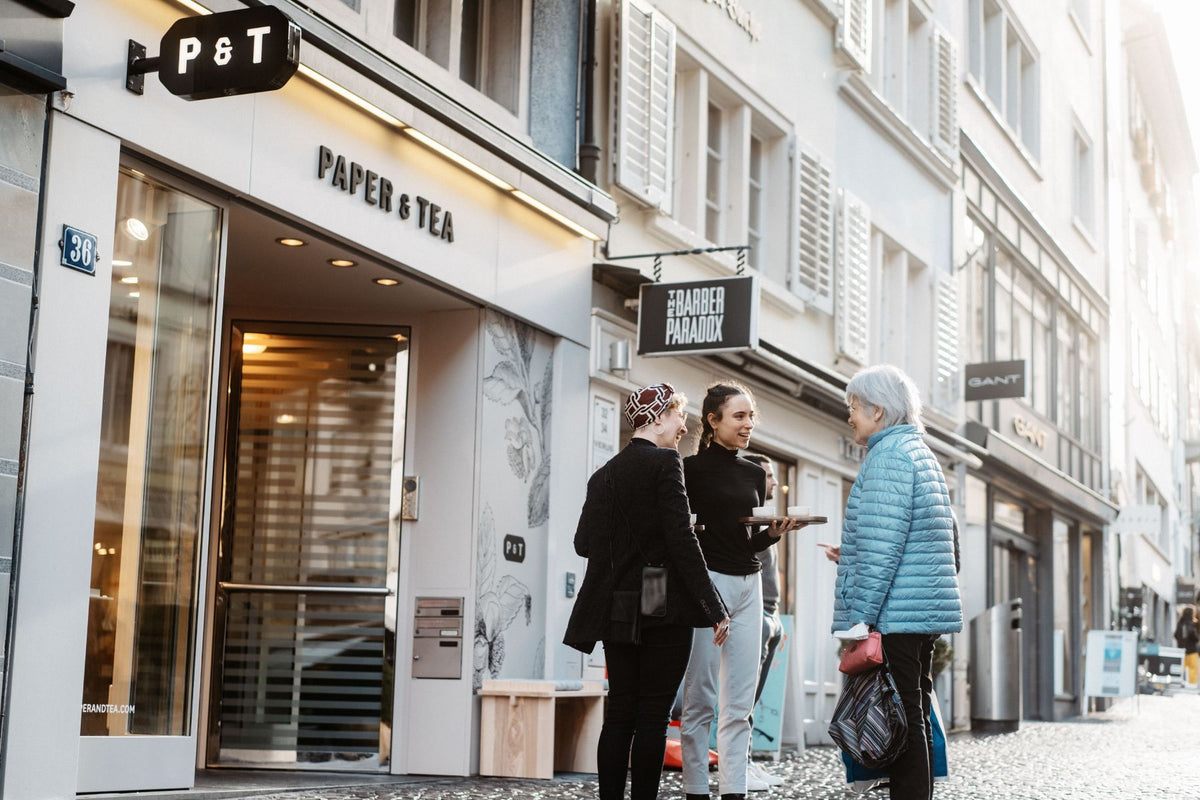 Straßenszene mit dem Teegeschäft P&T in Nummer 36, drei Frauen im Gespräch vor dem modernen Glas-Eingang.