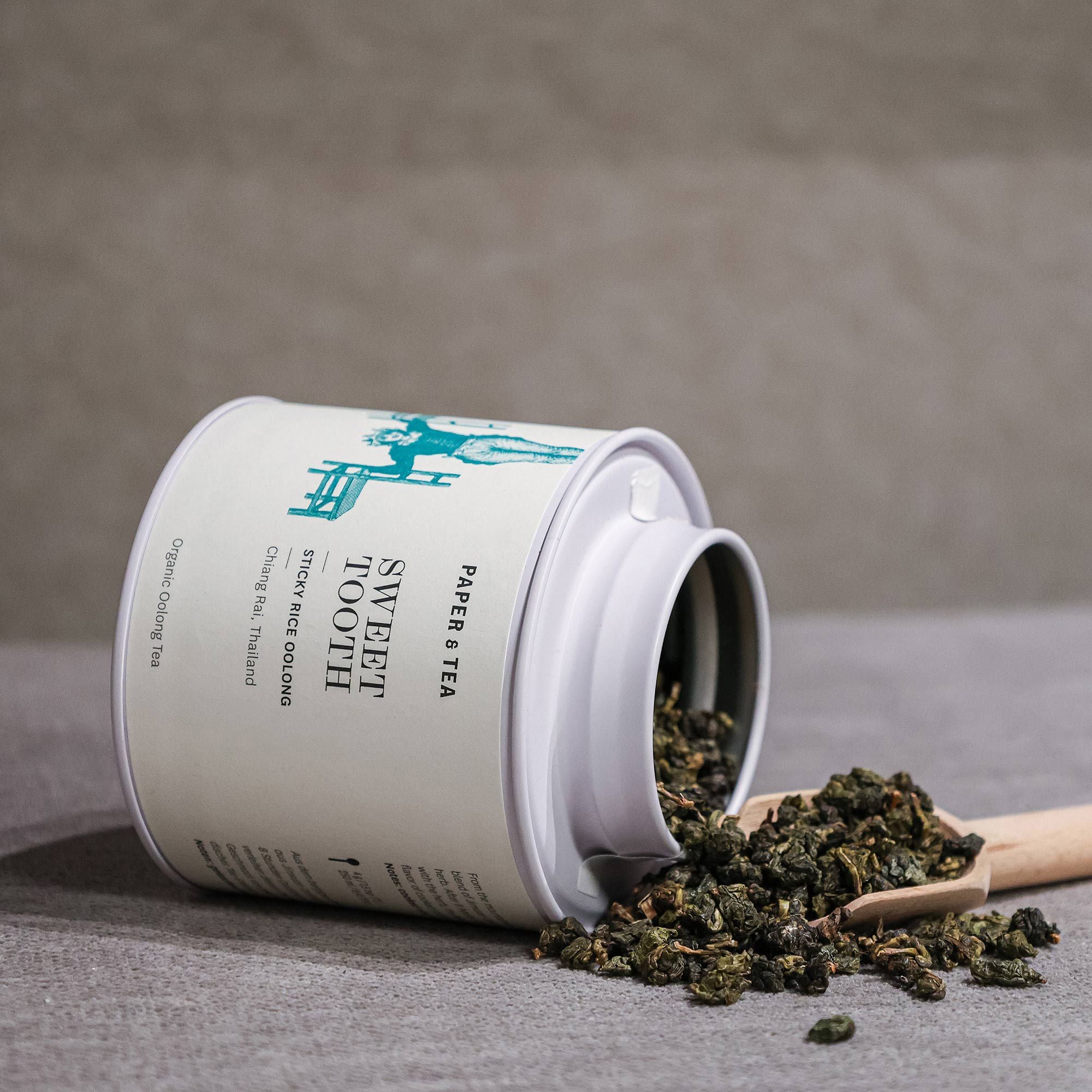 Weiße Teedose mit Aufschrift 'Sweet Tooth Sticky Rice Oolong', loser Bio-Oolong-Tee, Holzlöffel auf Stoff
