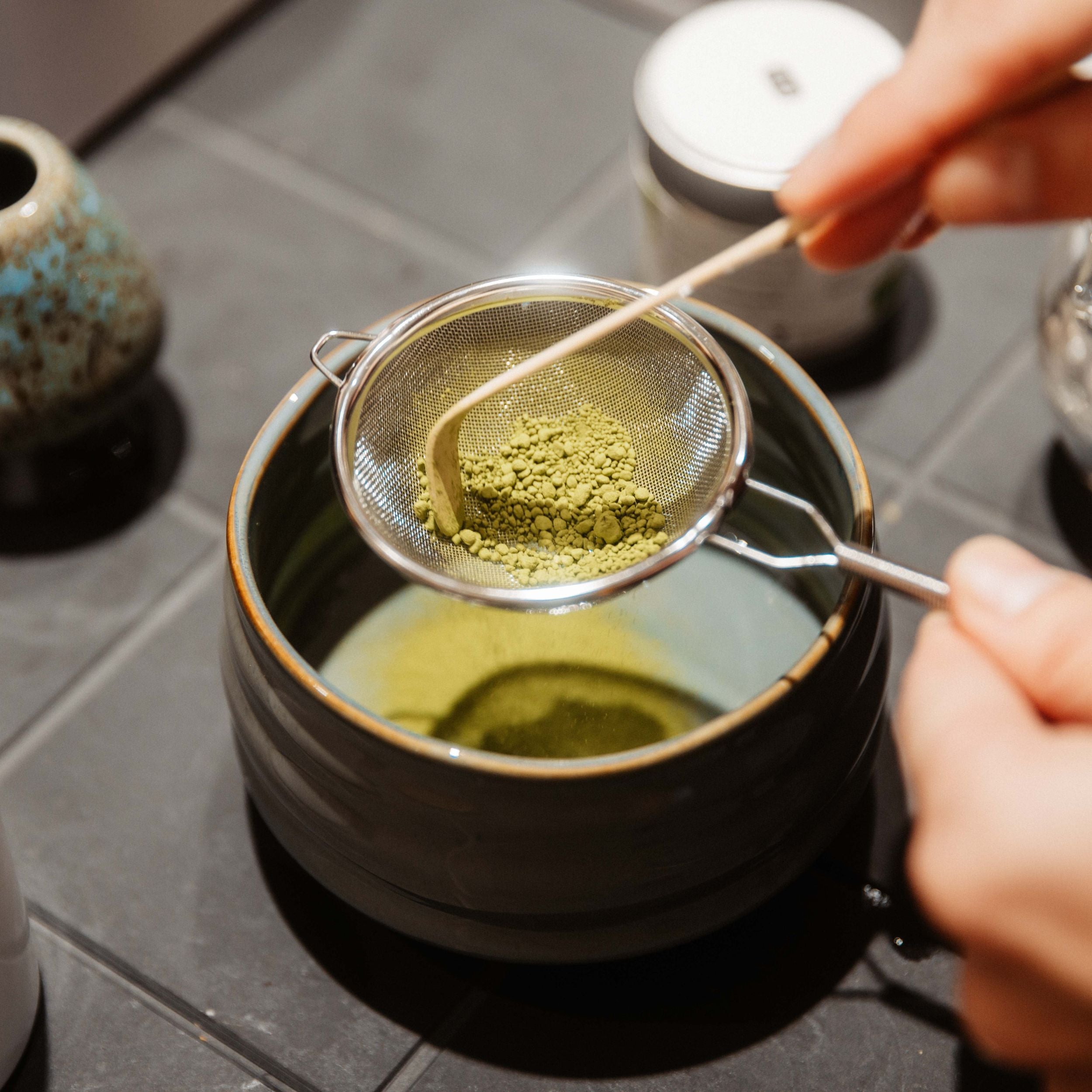 Hand siebt leuchtend grünes Matcha-Pulver durch ein feines Metallsieb in eine Keramikschale auf dunkler Fliese.